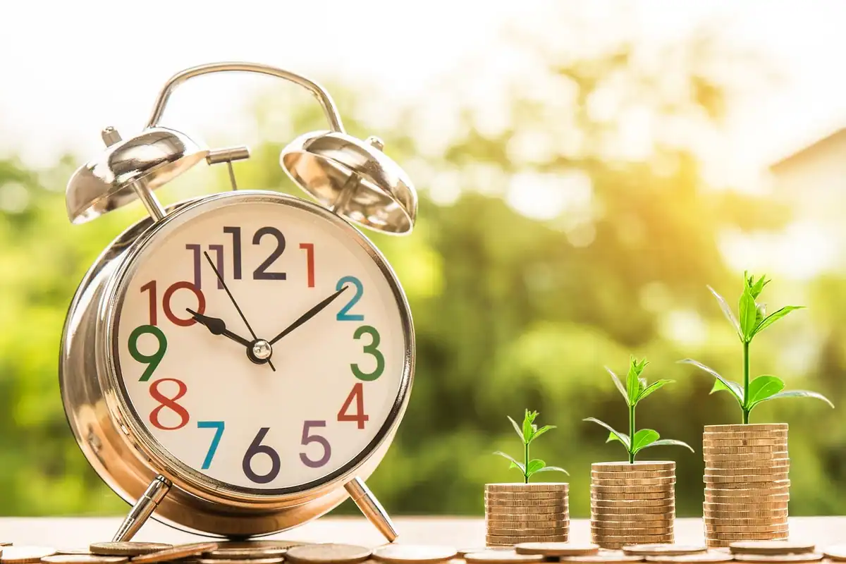 Ceas deșteptător lângă stive de monede din care cresc plante mici, simbol al economisirii pentru avans