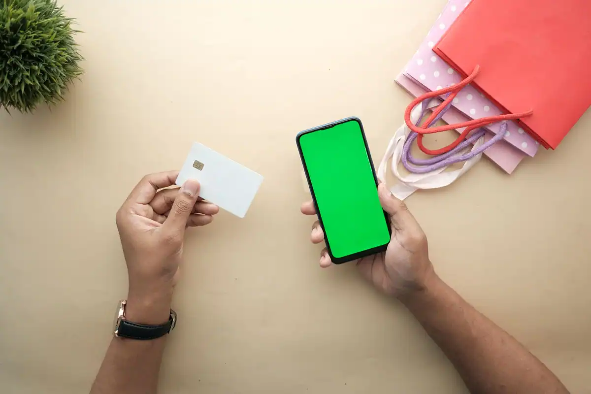 Mână care ține un card alb fără inscripții și un telefon, lângă pungi de cumpărături