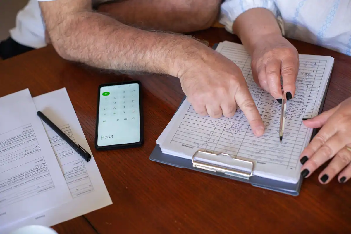Mâini care arată un rând dintr-un formular pe un clipboard, lângă un telefon cu calculator