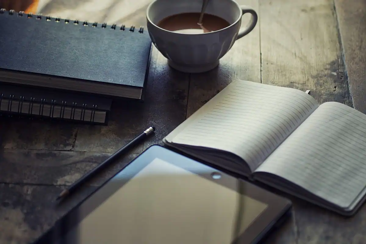 Un jurnal deschis, o cană de cafea și o tabletă pe un birou, într-un spațiu de lucru
