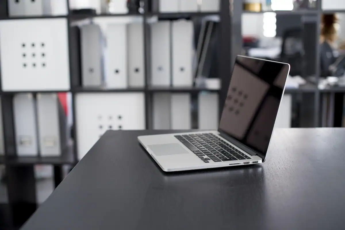 Laptop deschis pe un birou negru în fața unor rafturi cu bibliorafturi, pentru verificarea interogărilor